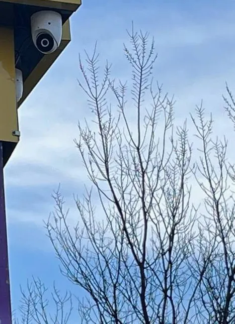 Device on a storefront fascia corner — reference photo from another AESS job, Sonos installation Upper West Side, Manhattan Upper West Side, Manhattan Sonos reference photo: small white dome device fixed under the corner of a yellow storefront fascia, blue sky and bare branches beyond
