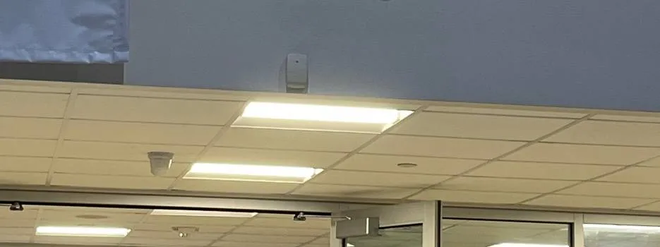Lit panels over glass doors below a grey wall — AESS reference photo, Sonos Upper West Side, Manhattan Upper West Side, Manhattan Sonos reference photo: lit light panels in a drop ceiling below a grey wall, with glass doors and a small round ceiling device beneath