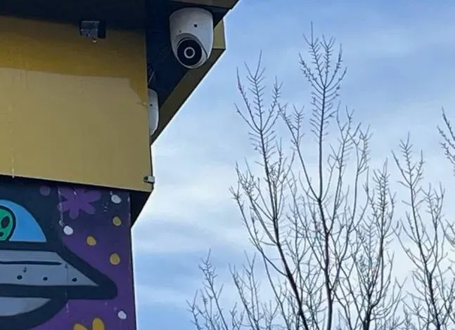 White turret camera under a fascia against bare winter trees β Putnam County Ubiquiti installation example Putnam County Ubiquiti camera installation example photo: a white turret camera mounted under a yellow fascia board beside bare winter tree branches and a blue sky