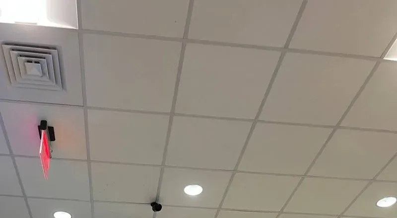 Small black camera on a tiled ceiling near a lit exit sign â Manhattan Uniview reference Manhattan Uniview installation reference photo: a small black camera on a white tiled ceiling near a lit red exit sign, an air diffuser and recessed lights
