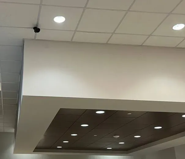 Small black camera above a showroom soffit with recessed lights â Manhattan Uniview reference Manhattan Uniview installation reference photo: a small black camera on a white drop ceiling above a dropped soffit with dark wood-tone panels and rows of recessed lights