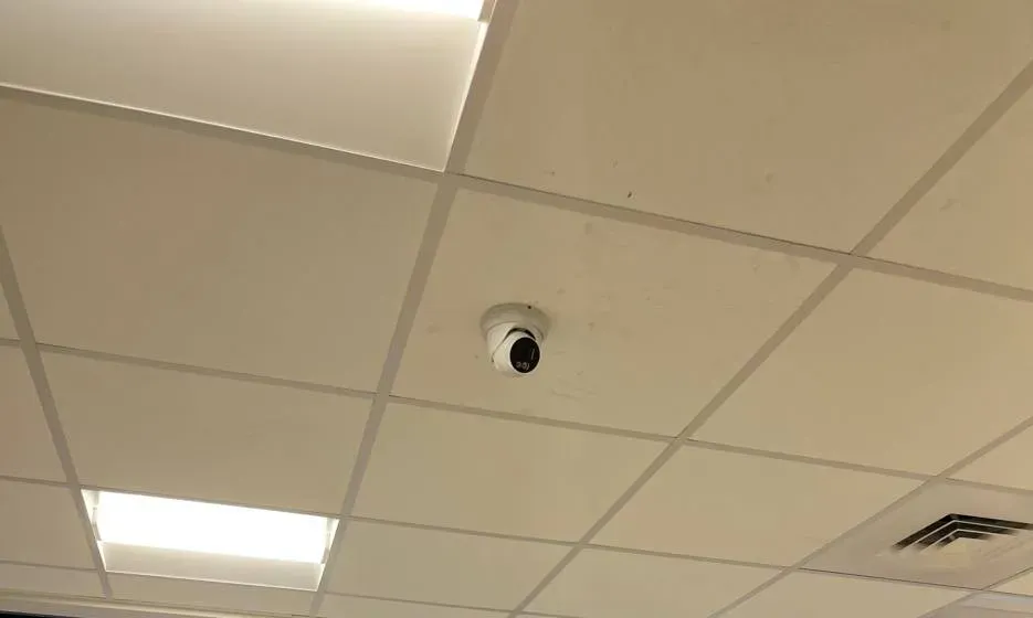 White turret camera on a ceiling tile between light panels — Uniview installation Nassau County example Uniview camera installation Nassau County example photo: a white turret camera on a ceiling tile between two light panels, with a return-air vent in the corner