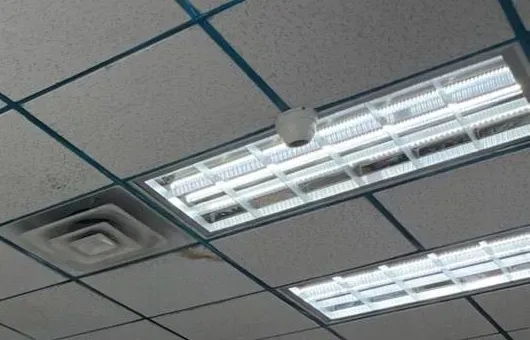 Mini dome beside a louvered light and an air diffuser — Staten Island Uniview install reference Staten Island Uniview install reference photo: a small white mini dome camera on a dark-grid drop ceiling next to a louvered light fixture and a square air diffuser