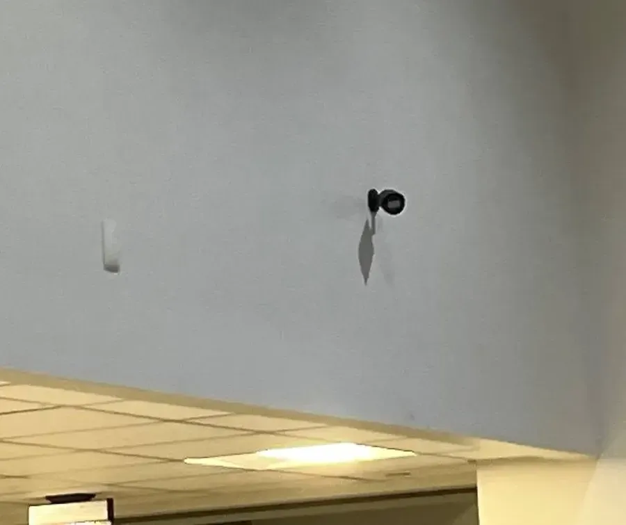 Small black camera high on a wall above a lowered ceiling — Staten Island Uniview shop interior reference Staten Island Uniview shop reference photo: a small black camera mounted high on a pale wall above a lowered ceiling section, casting a shadow, beside a small white sensor