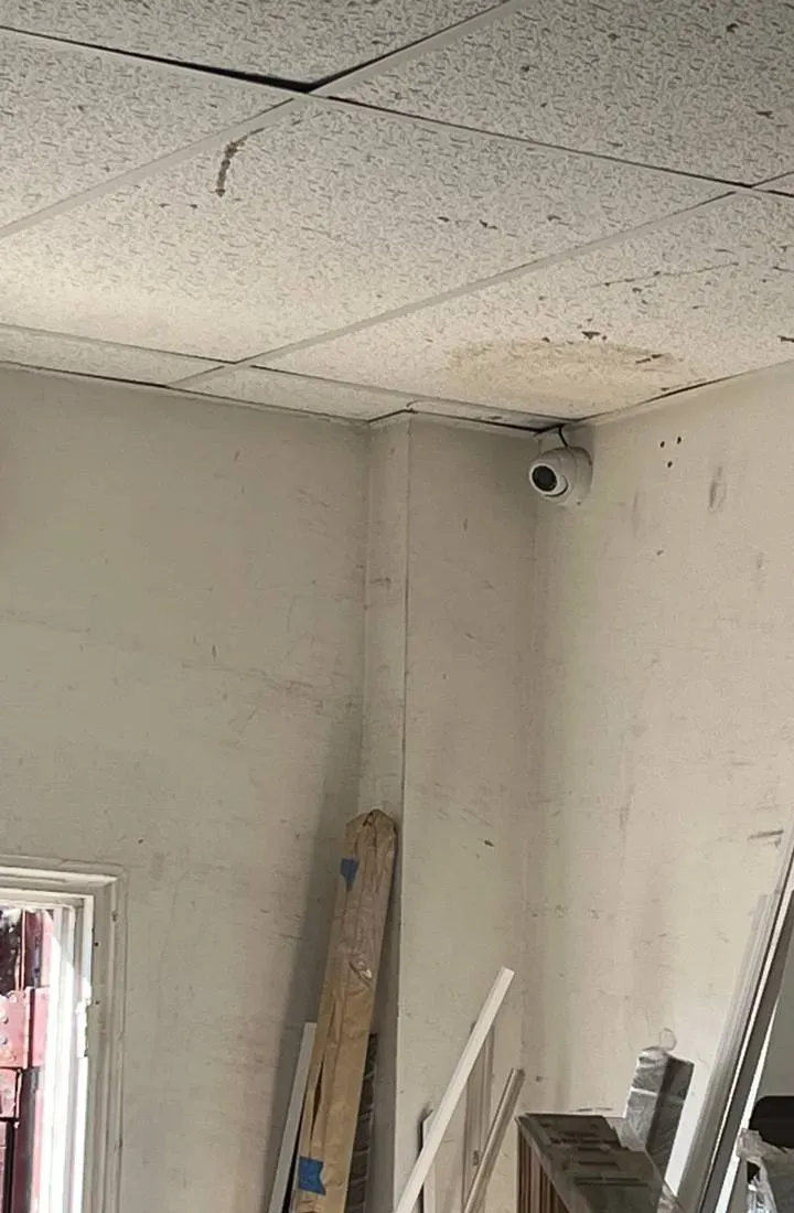 Turret camera high in a storage-room corner above stored boards — Staten Island Uniview basement reference Staten Island Uniview basement reference photo: a white turret camera high in the corner of a storage room under stained ceiling tiles, with boards leaning against the wall below