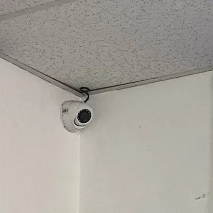 White dome in a ceiling corner with its cable looped above — Staten Island Uniview service reference Staten Island Uniview repair reference photo: a white dome camera fixed in the corner where a white wall meets a speckled ceiling, its cable looped above the housing