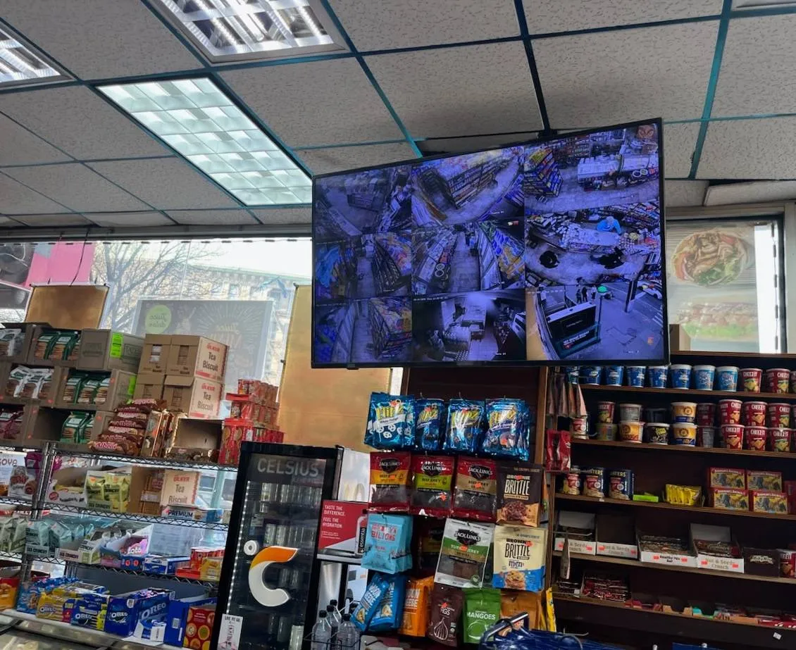 Video wall installation Long Island example photo: a large display hung from a drop ceiling above shop shelving and a drinks cooler, showing a grid of camera views