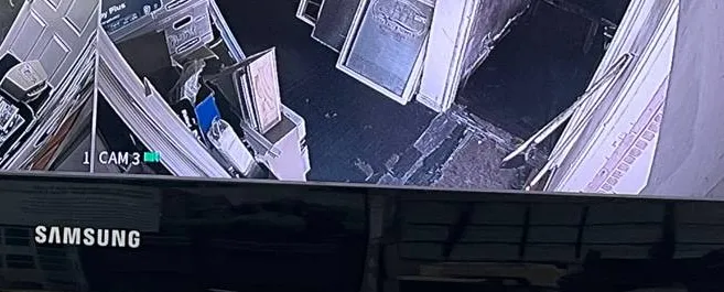 Video wall installation Long Island example photo: the glossy black lower bezel of a Samsung monitor below a camera tile showing a storeroom with stacked frames and an open door