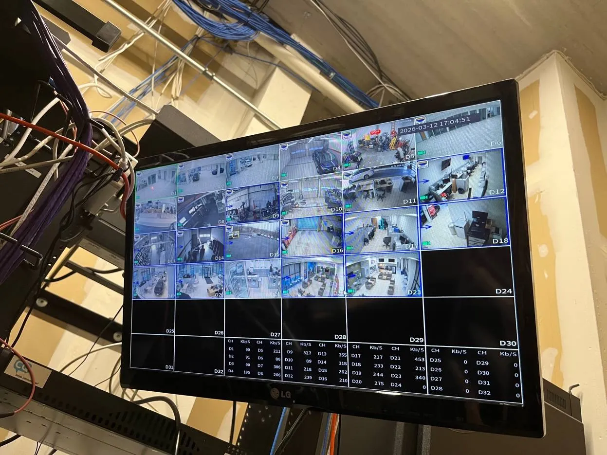 Video wall installation Long Island example photo: a single LG monitor showing a 32-tile grid of camera views and a per-channel bitrate table, hung beside an equipment rack with bundles of patch cables