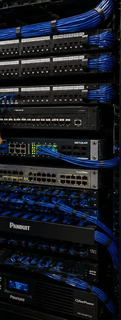 Equipment rack with battery backup at its base — Putnam County video wall power reference Putnam County video wall installation reference photo: a tall black equipment rack with patch panels, network switches and blue patch cables, and battery backup units at the bottom