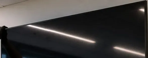 Gloved hands at the top edge of a wall display — Putnam County mounting reference Putnam County video wall installation reference photo: gloved hands hold the top edge of a large black wall display under white ceiling strip lights