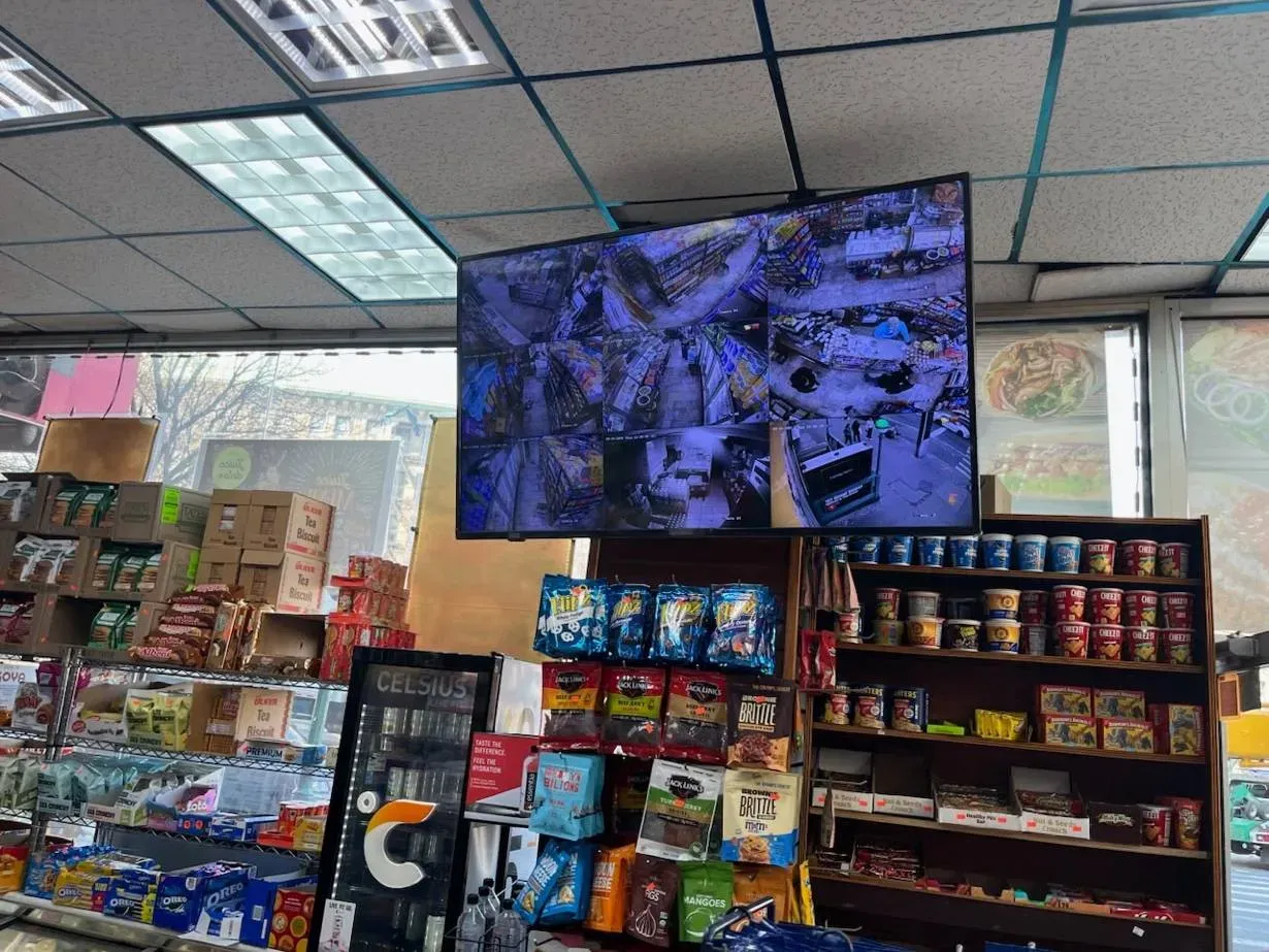 Ceiling-hung display above store shelving — Westchester storefront reference Westchester County video wall reference photo: a display hung from a drop ceiling above snack shelving in a small store, showing a grid of camera views, with the shop window and street behind