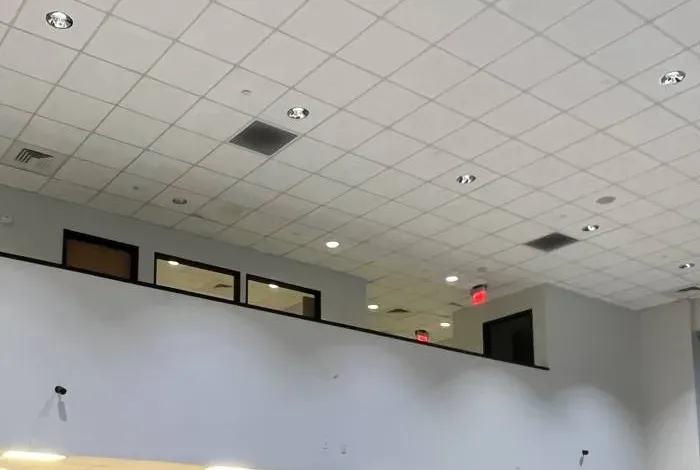 Tall white wall below mezzanine windows — Westchester double-height room reference Westchester County video wall reference photo: a tall white wall in a double-height room below a row of mezzanine windows, with a suspended tile ceiling, recessed lights and two small cameras on the wall