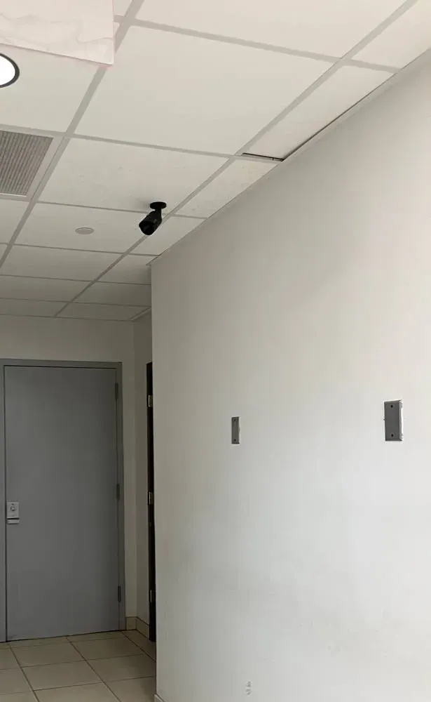 Wireless camera installation NYC reference photo: a black bullet camera on a short mount at the edge of a white drop-ceiling grid, above a grey hallway door, with plain white walls and two small metal wall plates
