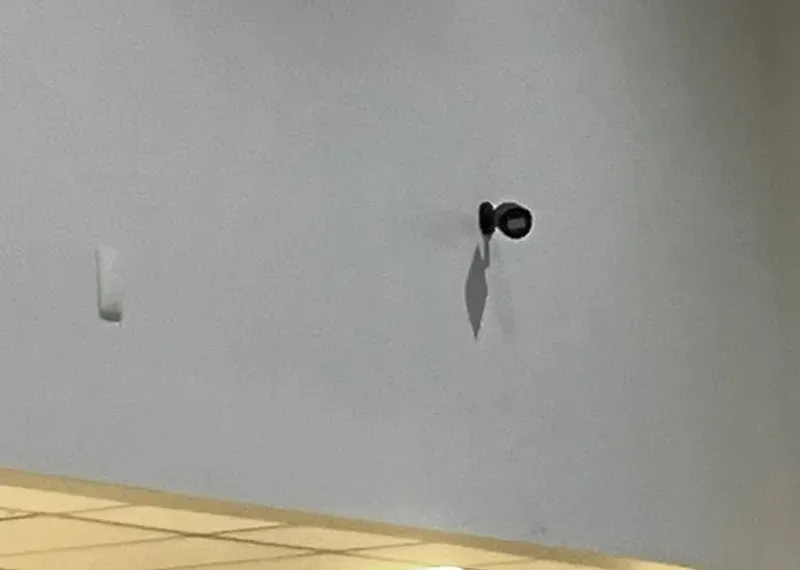 Wireless camera installation NYC reference photo: a small black camera on a short mount high on a bare pale wall, casting a long shadow below it, with a white wall plate to the left